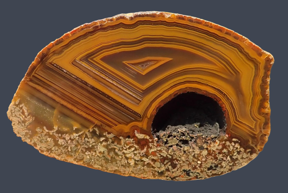Lilangabi agate 102 mm – full slab face, tight fortification banding sweeping into a sharp chevron point at centre, diagonal waterlines crossing the left side, amber, gold and deep brown, Lebombo Mountains