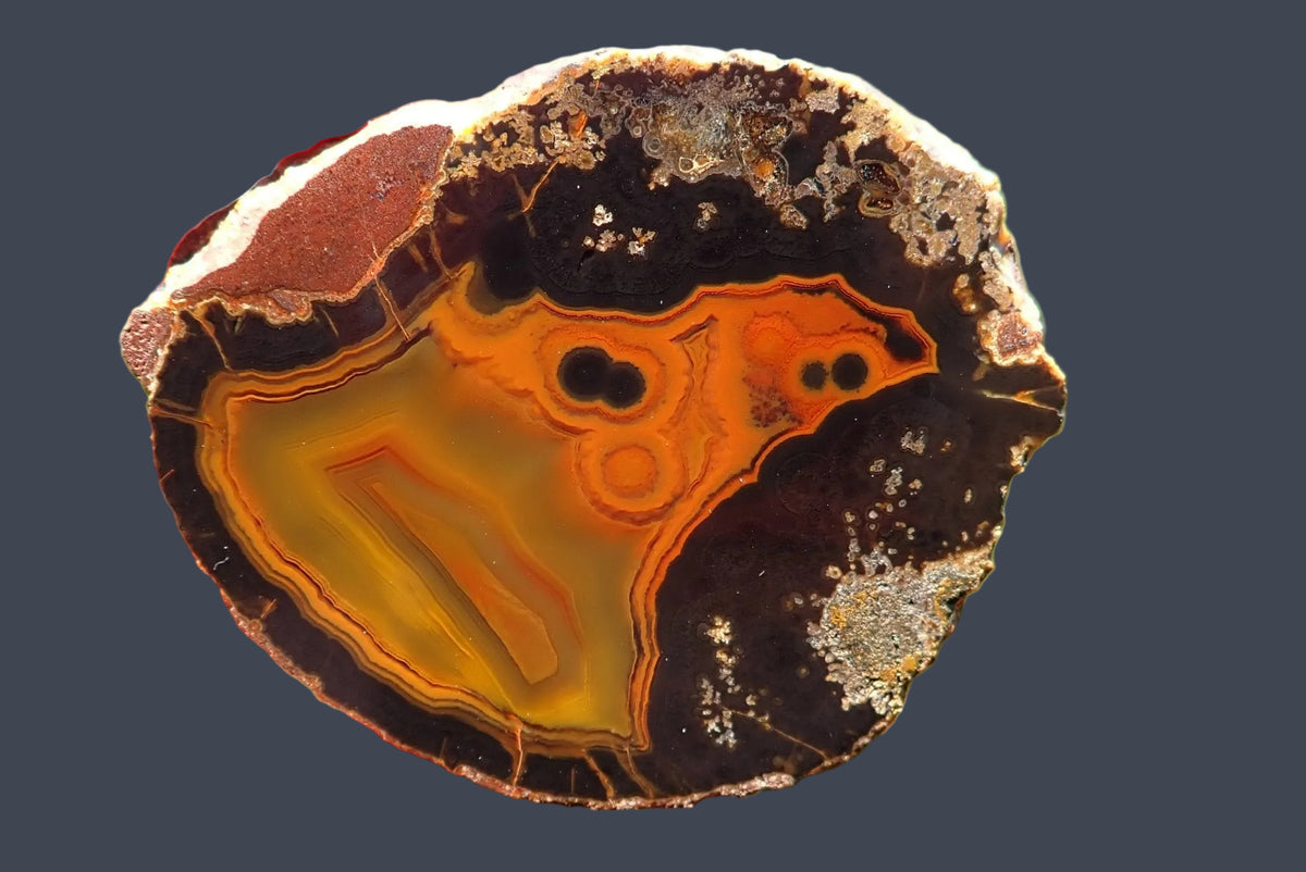 Lilangabi agate 63 mm – hand-lapped face with dark eyes, face-like pareidolia and fine fortification banding around an orange-gold centre, contrasting with a charcoal-brown surround.