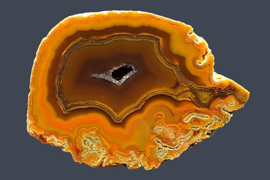 Lilangabi agate 71 mm – hand-lapped face with fine fortification banding around a brown centre, golden-orange chromatography, moss inclusions along the lower edge and a central drusy vug.