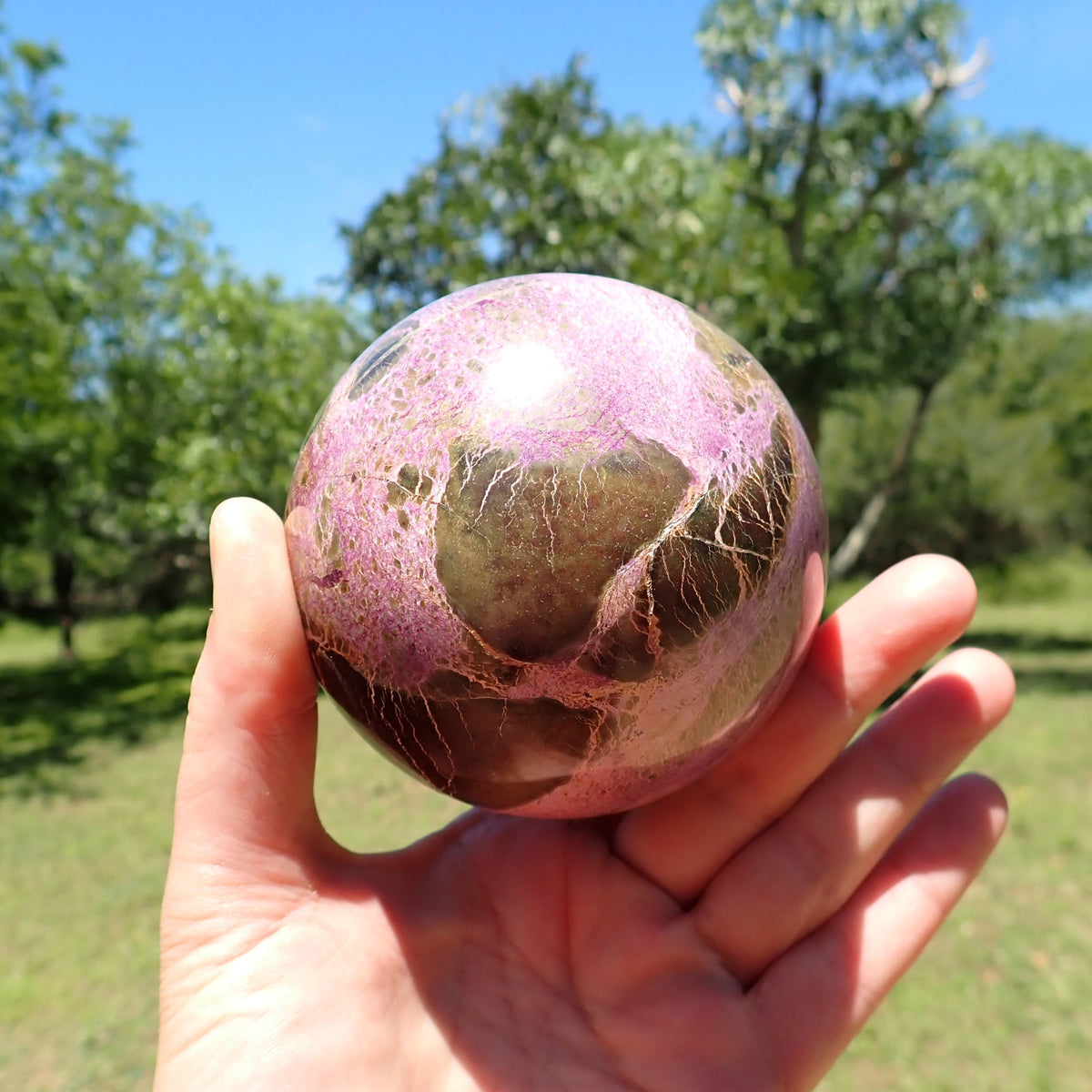 Stichtite with Serpentine sphere held in an open hand for scale, showing large brown patches within pink-purple intergrowths.