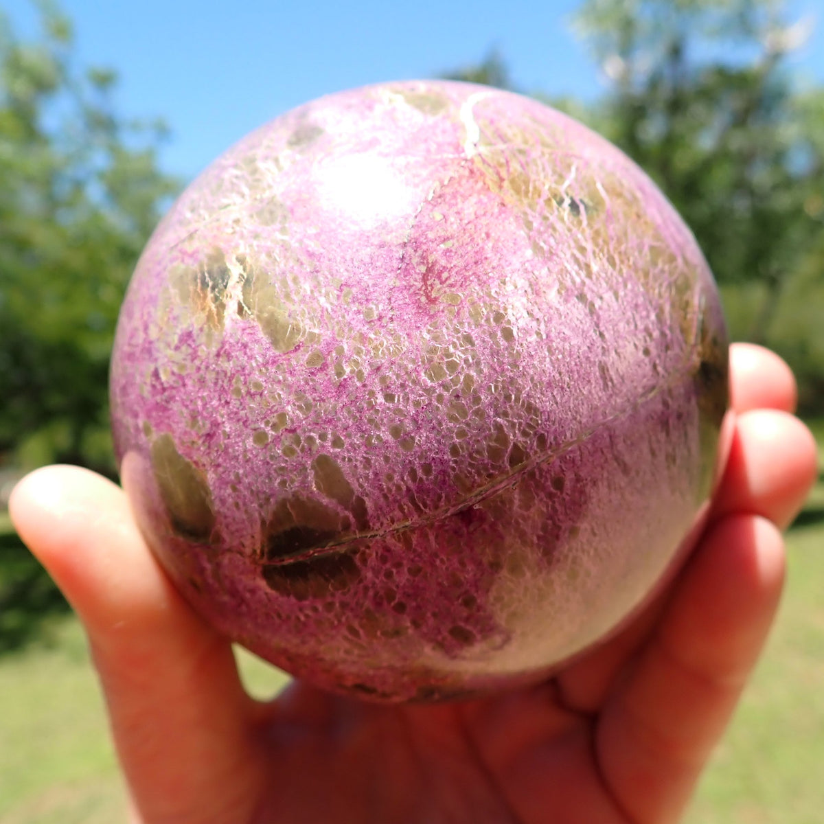 Stichtite with Serpentine sphere with pink-purple intergrowths surrounding scattered olive-brown patches, held in natural sunlight.